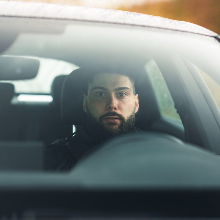 Begleitschutz Köln & deutschlandweit. Derlo CP Mann mit Bart sitzt am Steuer eines Autos und schaut in den Rückspiegel.